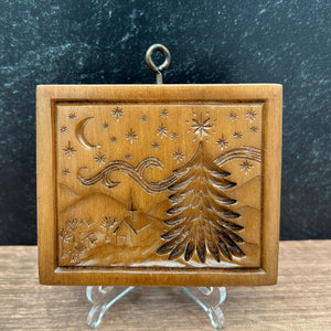 A square cookie mold featuring a stylized Christmas tree, stars, and crescent moon, displayed on a clear stand against a split dark gray and wood background.