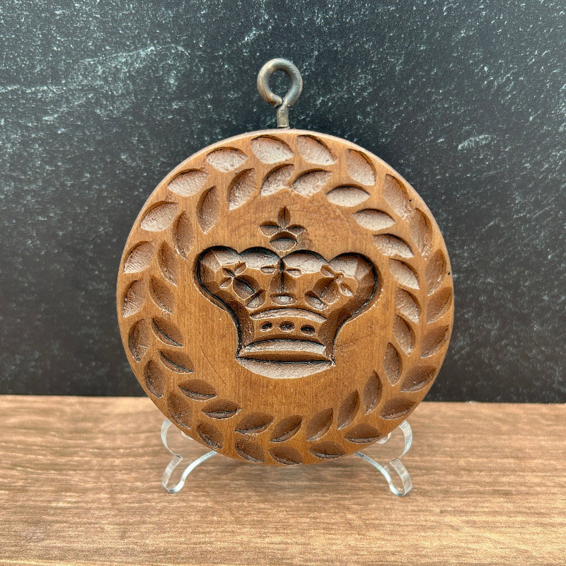 A round wooden cookie mold with an engraved crown and laurel wreath design, displayed on a small clear stand against a split dark gray and wood-grain background.