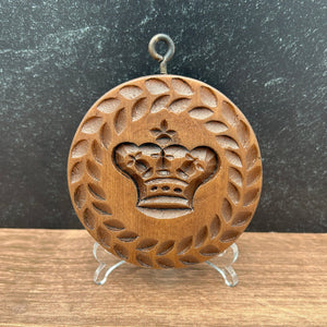A round wooden cookie mold with an engraved crown and laurel wreath design, displayed on a small clear stand against a split dark gray and wood-grain background.