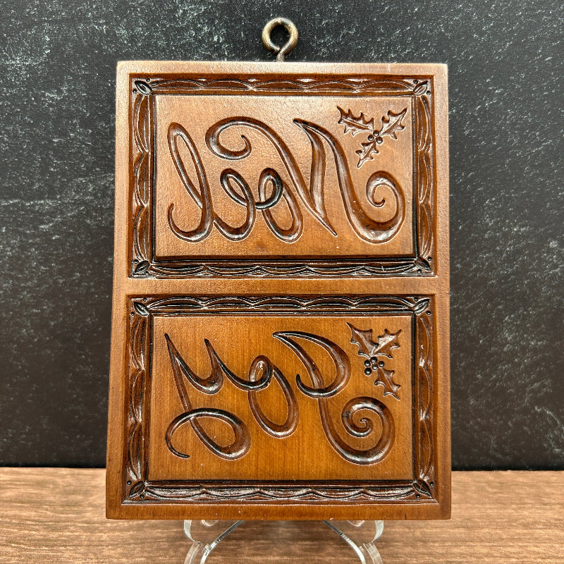 A rectangular cookie mold with two carved panels featuring stylized monogram-like lettering and holly leaves, displayed on small clear stands against a dark textured wall and wood surface.