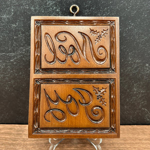 A rectangular cookie mold with two carved panels featuring stylized monogram-like lettering and holly leaves, displayed on small clear stands against a dark textured wall and wood surface.