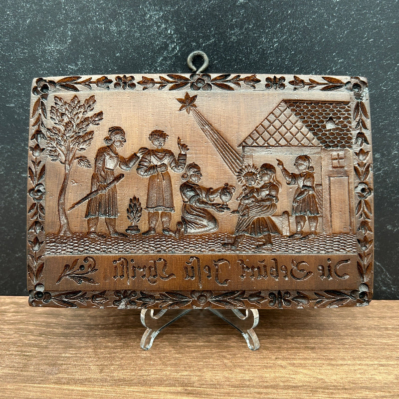 A rectangular carved cookie mold with a hinged metal clasp and hanging loop, featuring embossed religious or historical figures and text, photographed on a split dark gray and wood surface.