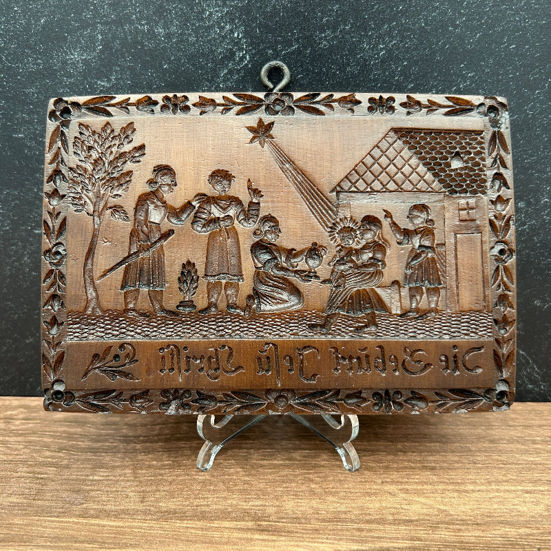 A rectangular carved cookie mold with a hinged metal clasp and hanging loop, featuring embossed religious or historical figures and text, photographed on a split dark gray and wood surface.