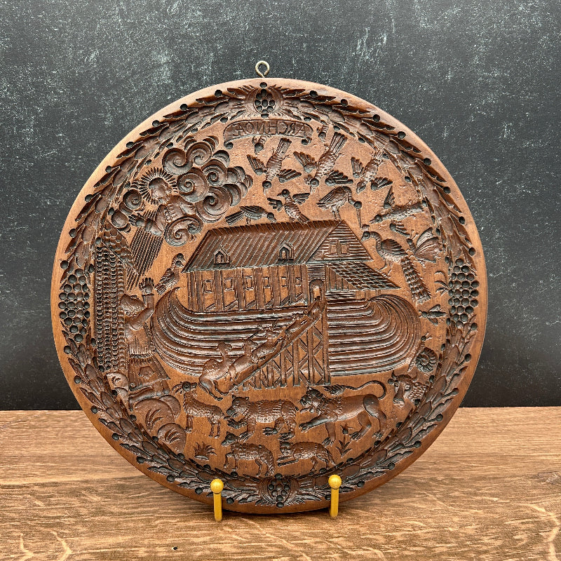 A round, hand-carved wooden Noah's Ark wall plaque displayed on a wooden stand against a dark textured wall.