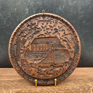 A round, hand-carved wooden Noah's Ark wall plaque displayed on a wooden stand against a dark textured wall.