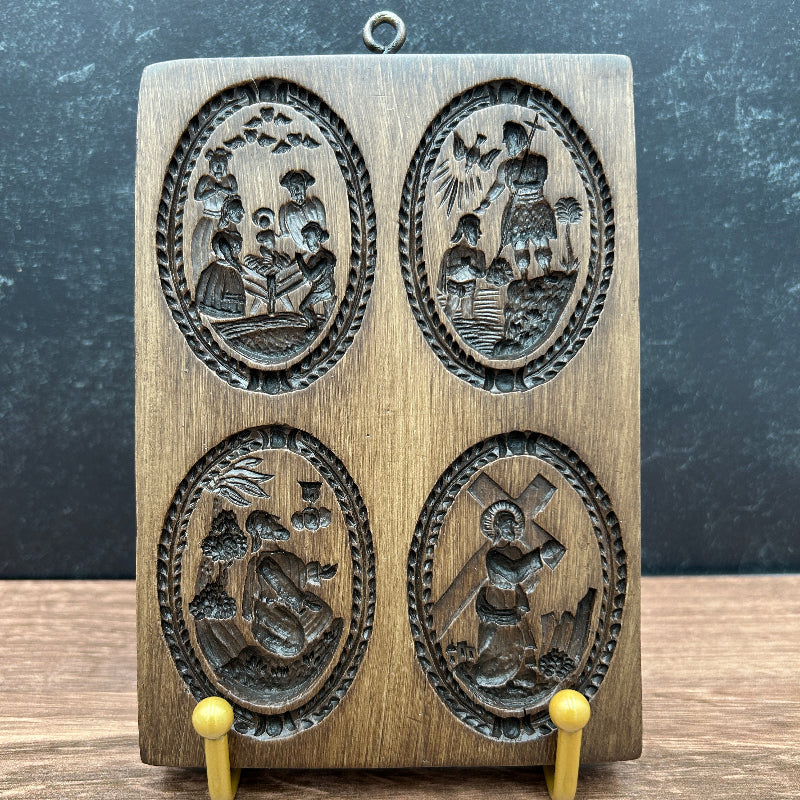A rectangular wooden carving mold or plaque with four oval engraved scenes, displayed upright on small yellow stands against a dark textured background and wood surface.