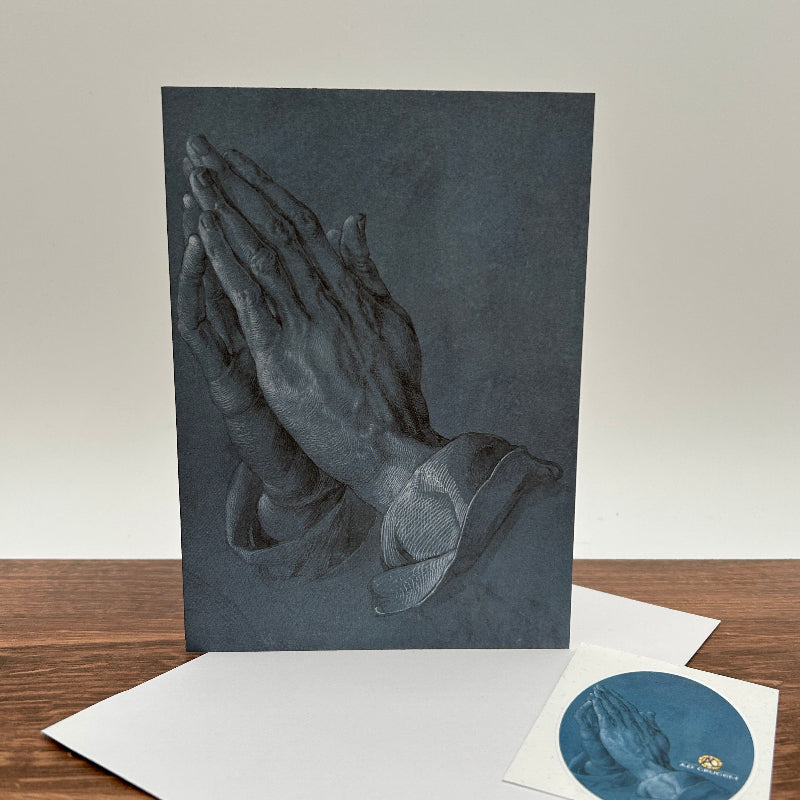 A dark slate greeting card of praying hands displayed on a wooden table with a white envelope and a small circular sticker.