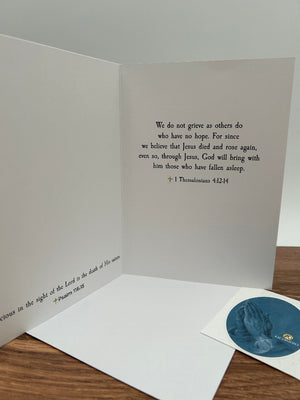An open white prayer card or booklet on a wooden surface, showing printed Bible verses on the inside pages, with a small oval blue prayer card placed beside it.